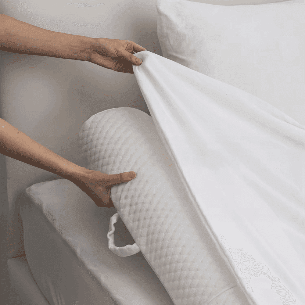 Hands fitting the white sheet over a quilted foam bolster at the edge of the bed