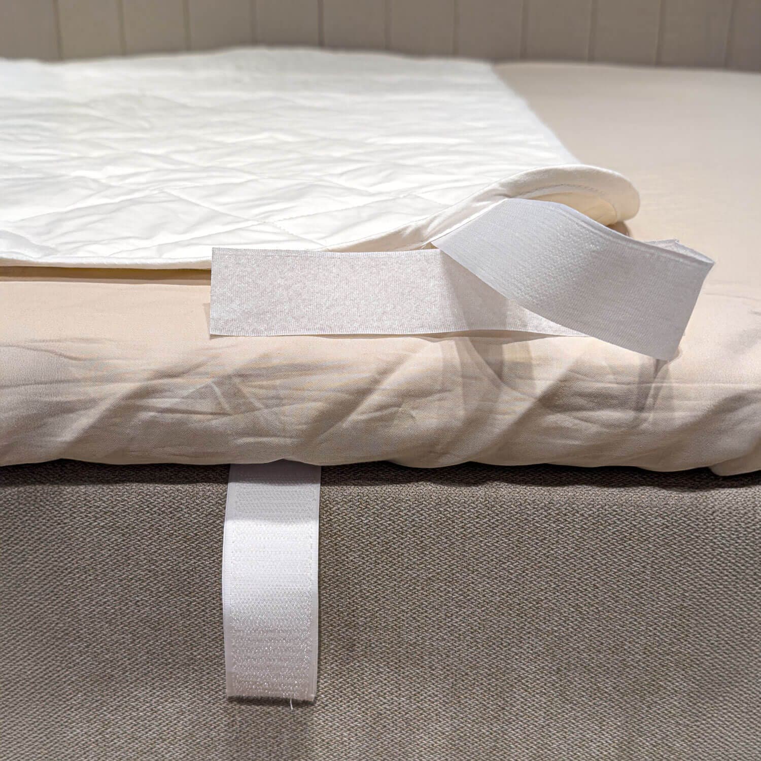Corner of the white quilted mattress protector folded back to reveal the hook-and-loop strap around the edge of the bed