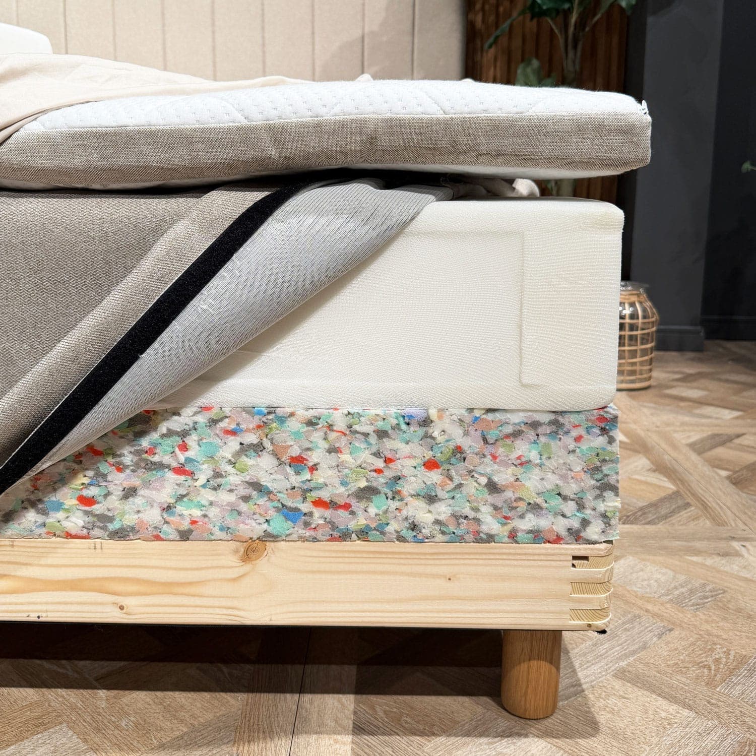 Bed layers opened at the corner, showing the mattress topper, sprung mattress with its cover opened and the bed base's foam edge above the wooden frame