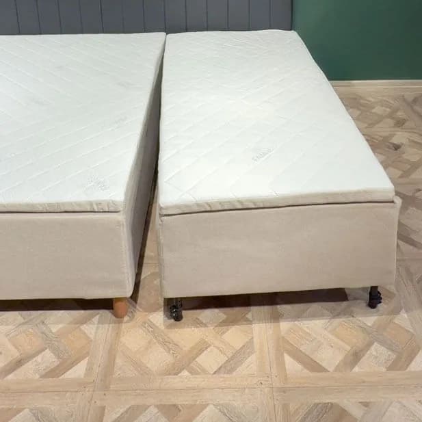 Two fully assembled bed modules stand side by side on a light wood floor, with a narrow gap between them. Each module has its own beige cover, quilted mattress and legs.